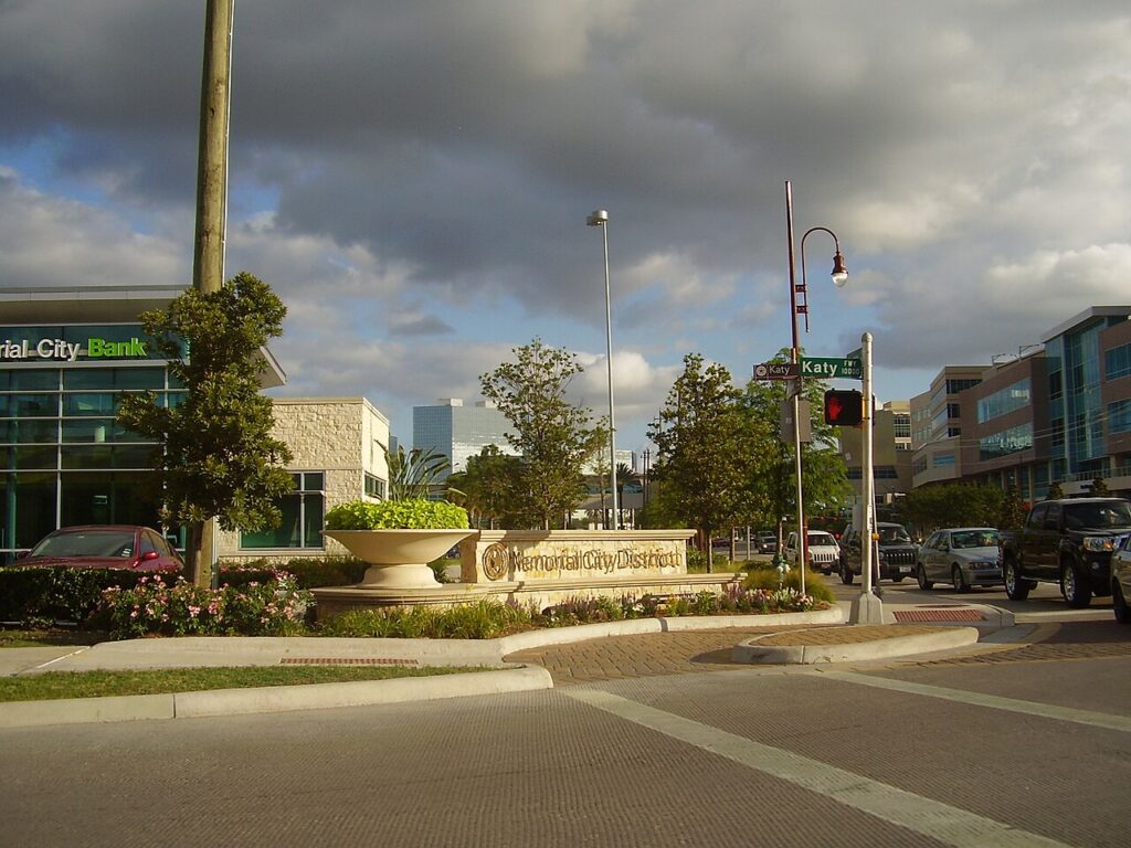 Street-level view of the Memorial City District sign at the intersection of Katy Freeway in Houston, Texas, featuring a decorative planter, landscaped median, commercial buildings, and overcast skies.